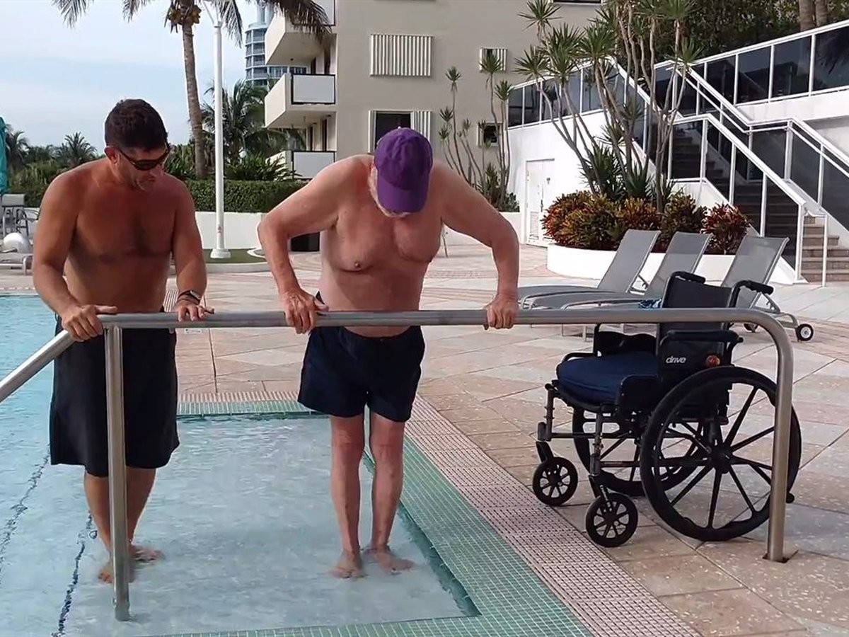 A trainer steadying a client on the pool-entry handrail, with a wheelchair waiting on the deck behind them