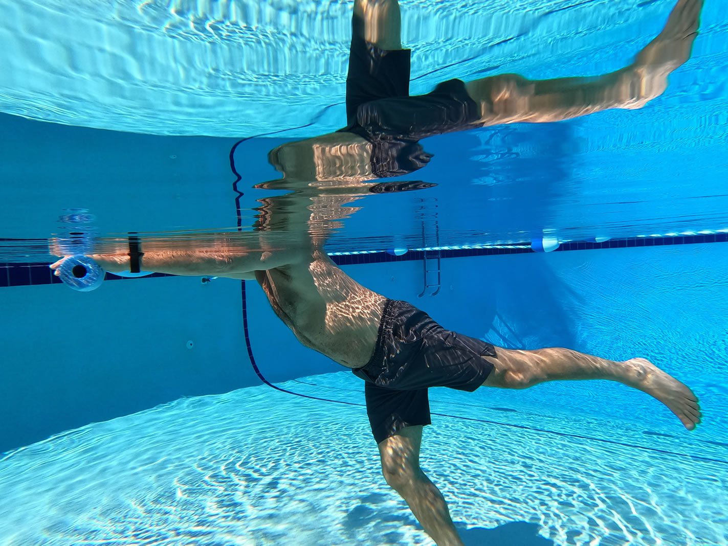 Underwater side view of Stuart Davidson performing an upright aquatic leg movement with a clear reflection at the water surface.
