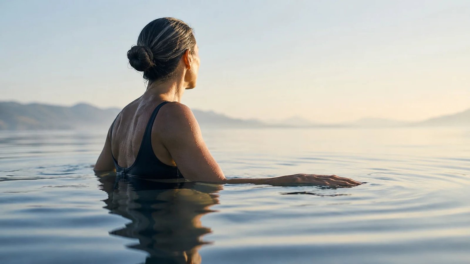 A woman immersed to chest depth in calm open water, seen from behind at a three-quarter angle facing right, one arm extended along the surface, distant mountains at the horizon.