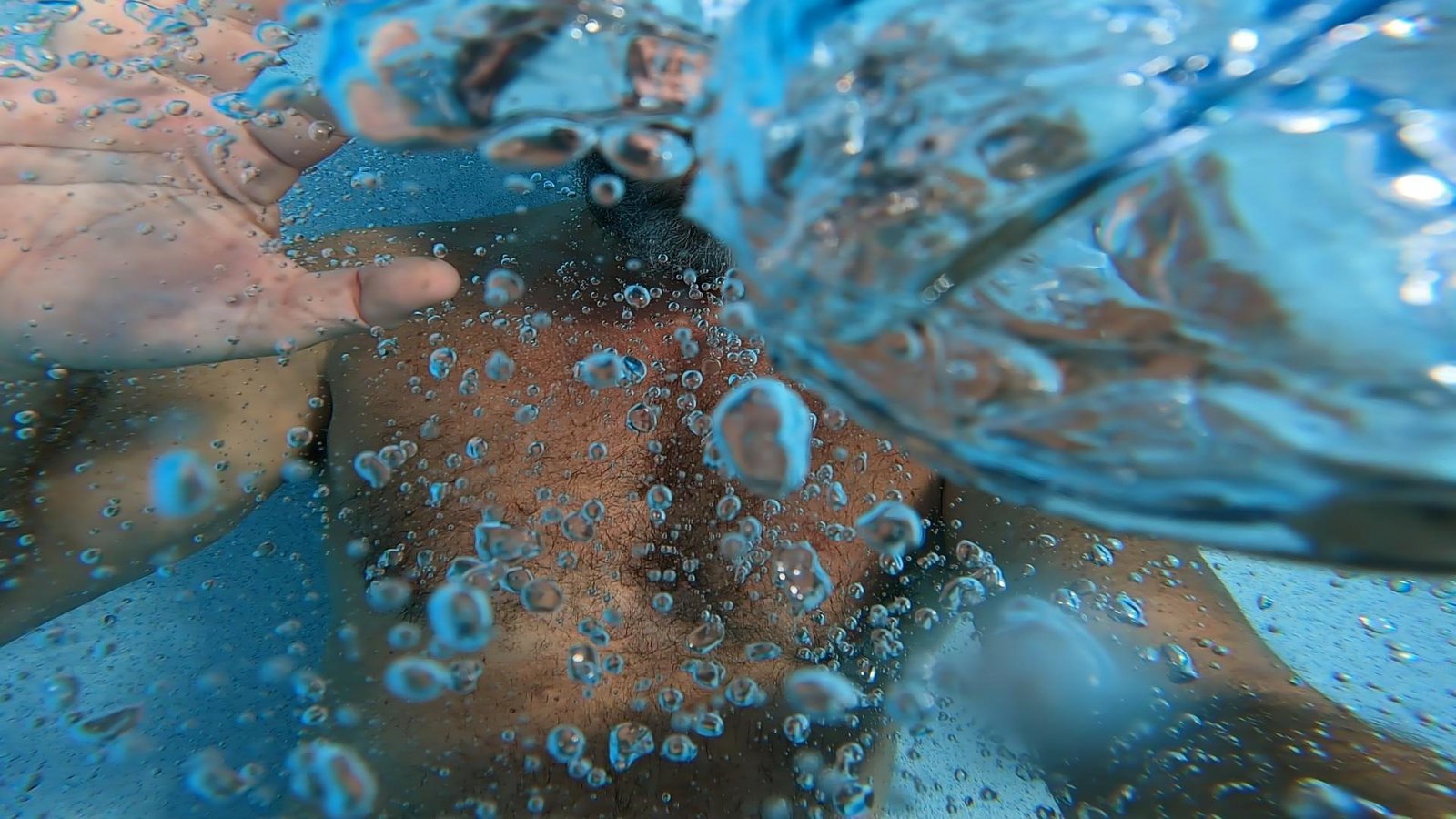 Air bubbles clinging to the skin of a man's chest just below the surface, one open hand raised toward the camera, the broken water surface silvered with light overhead.