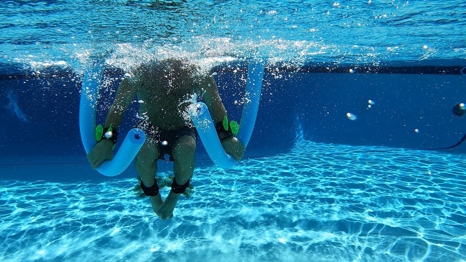 Athlete suspended underwater mid-exercise, driving two buoyant foam noodles down against the water's resistance